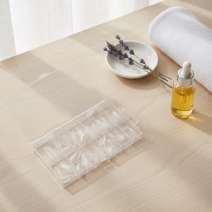 May include: A clear plastic case filled with oval-shaped artificial nails. A small white dish with lavender sprigs, a bottle of oil, and a rolled white towel are also on the light wood surface.