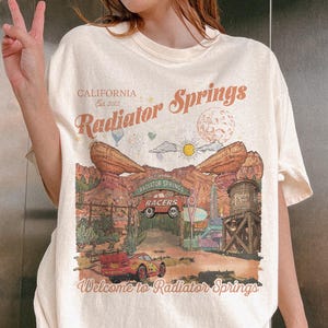K&ouml;nnte beinhalten: Ein cremefarbenes Kurzarm-T-Shirt mit einem Grafikdruck von Radiator Springs in Kalifornien. Das Design zeigt W&uuml;stenfelsformationen, ein rotes Rennauto, einen Wasserturm sowie den Schriftzug "Radiator Springs" und "Welcome to Radiator Springs".