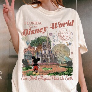 K&ouml;nnte beinhalten: Ein cremefarbenes T-Shirt mit einem Grafikdruck von Florida Disney World, einschlie&szlig;lich Schloss, Epcot-Sph&auml;re und Mickey Mouse. Der Text lautet Florida Est. 1971 Disney World und The Most Magical Place On Earth in rotbrauner Schrift.