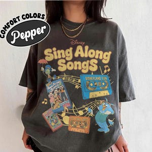 May include: A charcoal gray short-sleeve t-shirt featuring a graphic print with the text Disney Sing Along Songs. The design includes illustrations of Jiminy Cricket, a blue Disneyland Fun cassette tape, and VHS covers with musical notes.