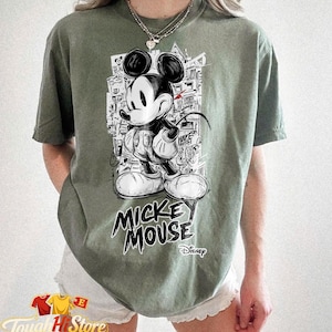May include: Sage green t-shirt with a black and white Mickey Mouse graphic. The design includes the text "Mickey Mouse" in a bold font, with the Disney logo. The shirt has a relaxed fit.