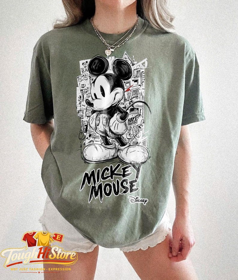 May include: Sage green t-shirt with a black and white Mickey Mouse graphic. The design includes the text "Mickey Mouse" in a bold font, with the Disney logo. The shirt has a relaxed fit.