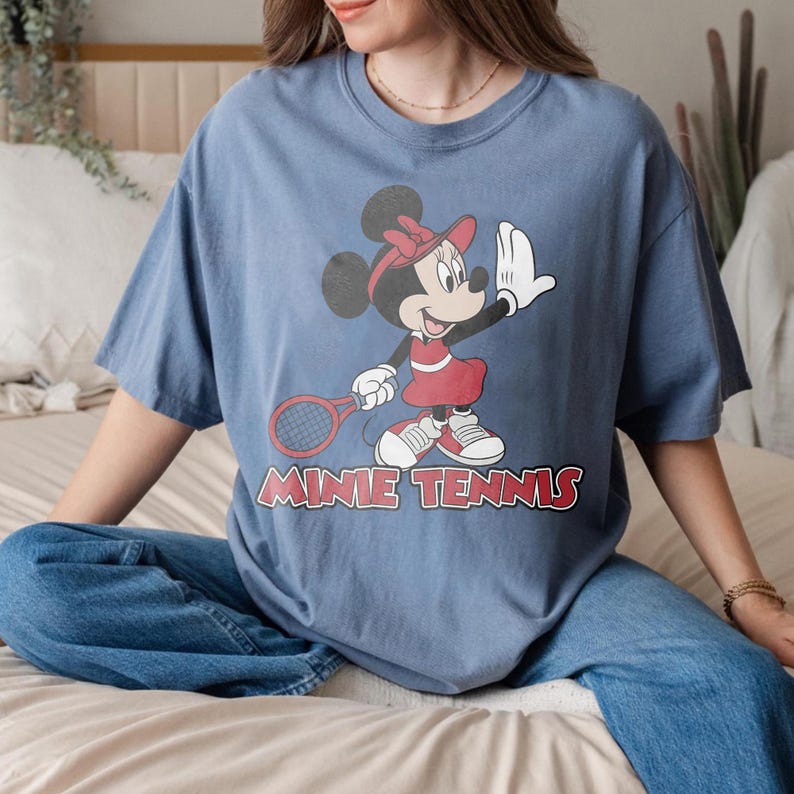 以下が含まれることがあります： テニスをするミニーマウスのグラフィックがプリントされた青いTシャツ。ラケットを持ち、手を振っています。「MINNIE TENNIS」の文字が赤でプリントされています。ゆったりとしたフィット感で、カジュアルなスタイルです。