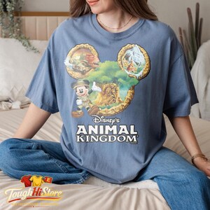 May include: A blue t-shirt featuring a Mickey Mouse design with the text "Disney's Animal Kingdom." The design includes images of a safari vehicle, mountains, and a tree with Mickey Mouse in a safari outfit. The shirt is a relaxed fit.