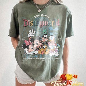 Puede incluir: Camiseta verde oliva con el texto "Walt Disney's Disneyworld" y un gr&aacute;fico de personajes de Disney, incluyendo Mickey y Minnie Mouse. La camiseta tambi&eacute;n tiene el texto "est 1971" y "Where dreams come true."