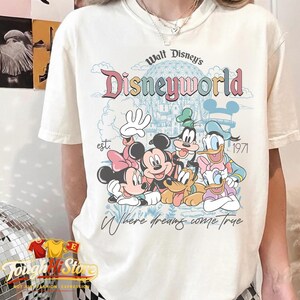 Puede incluir: Camiseta color crema con un gr&aacute;fico colorido de personajes de Disney, incluyendo Mickey y Minnie Mouse, Goofy, Donald y Daisy Duck. Tambi&eacute;n se muestra el texto "Walt Disney's Disneyworld" y "Where dreams come true".