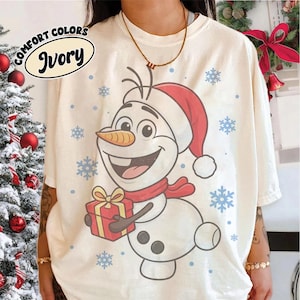 May include: An ivory t-shirt featuring a graphic of a smiling snowman wearing a red Santa hat and scarf, holding a red gift box with a yellow ribbon. The design is surrounded by blue snowflakes and includes the text COMFORT COLORS IVORY.