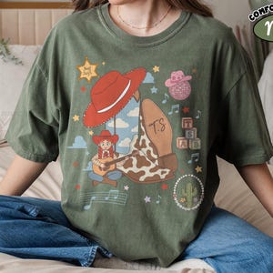May include: A moss green t-shirt featuring a graphic print of a cowgirl playing a guitar, a red cowboy hat, a cow-print boot with the initials TS, a pink disco ball hat, alphabet blocks, a cactus, musical notes, and a star with the text Feel Swift.
