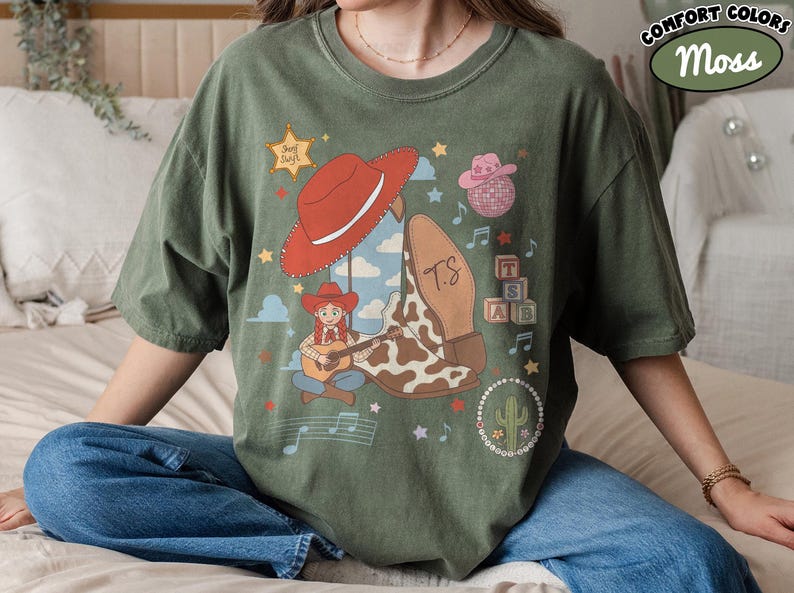 May include: A moss green t-shirt featuring a graphic print of a cowgirl playing a guitar, a red cowboy hat, a cow-print boot with the initials TS, a pink disco ball hat, alphabet blocks, a cactus, musical notes, and a star with the text Feel Swift.
