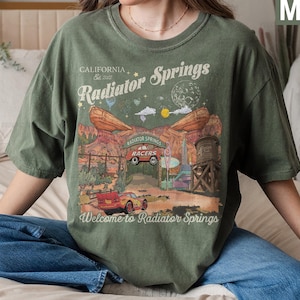 K&ouml;nnte beinhalten: Ein moosgr&uuml;nes Kurzarm-T-Shirt mit einem Grafikdruck von Radiator Springs, inklusive des Textes California Est 2022, Radiator Springs, Radiator Springs Racers und Welcome to Radiator Springs sowie einer Abbildung eines roten Rennwagens.