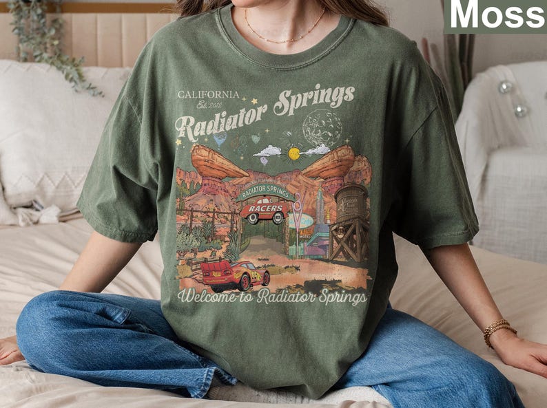 K&ouml;nnte beinhalten: Ein moosgr&uuml;nes Kurzarm-T-Shirt mit einem Grafikdruck von Radiator Springs, inklusive des Textes California Est 2022, Radiator Springs, Radiator Springs Racers und Welcome to Radiator Springs sowie einer Abbildung eines roten Rennwagens.