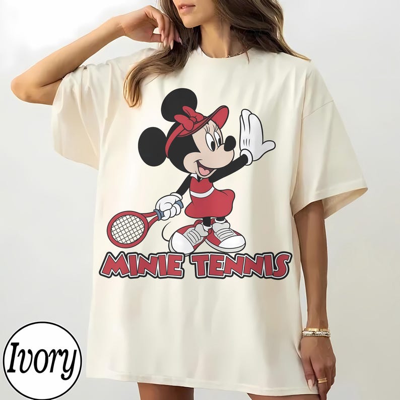 以下が含まれることがあります： アイボリー色のTシャツ。赤いテニスウェアを着たミニーマウスの漫画が、ラケットを持ち手を振っています。「MINNIE TENNIS」の文字が赤色でプリントされています。ゆったりとしたフィット感のシャツです。