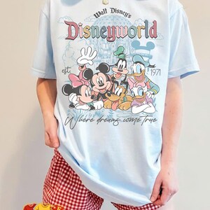 Puede incluir: Camiseta azul claro con un dise&ntilde;o de Disney con Mickey y Minnie Mouse, Goofy y Donald y Daisy Duck. La camiseta tiene el texto "Walt Disney's Disneyworld est. 1971" y "Where dreams come true."
