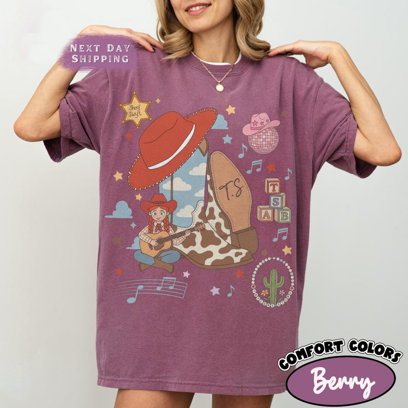 May include: A berry-colored t-shirt featuring a graphic print of a cowgirl playing a guitar, a large red cowboy hat, a cow-print boot with the initials TS, a disco ball, alphabet blocks, a cactus, stars, and musical notes on the front.