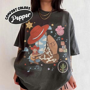 May include: A charcoal gray t-shirt featuring a graphic print with a red cowboy hat, a cow-print boot, a character playing a guitar, a pink disco ball, alphabet blocks with the letters T, S, and AB, a cactus, stars, and musical notes.