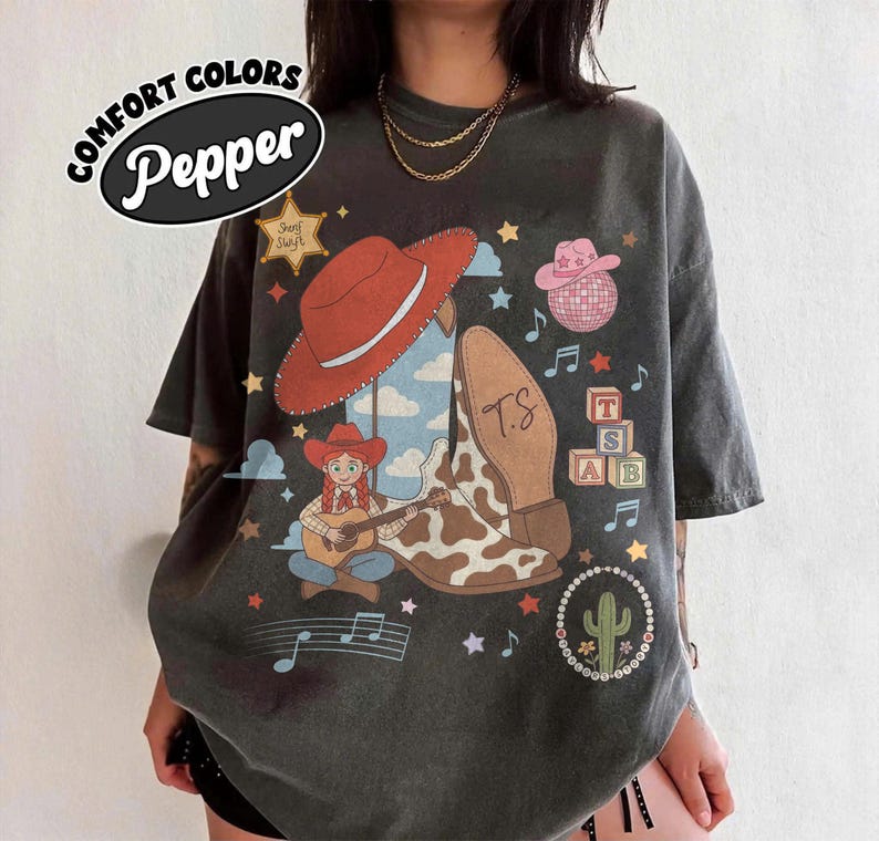 May include: A charcoal gray t-shirt featuring a graphic print with a red cowboy hat, a cow-print boot, a character playing a guitar, a pink disco ball, alphabet blocks with the letters T, S, and AB, a cactus, stars, and musical notes.