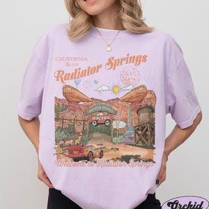 K&ouml;nnte beinhalten: Ein hellviolettes Kurzarm-T-Shirt mit einem Grafikdruck von Radiator Springs und dem Text California Est. 2012, Radiator Springs sowie Welcome to Radiator Springs. Das Design zeigt ein rotes Rennauto, W&uuml;stenfelsformationen und einen Wasserturm.