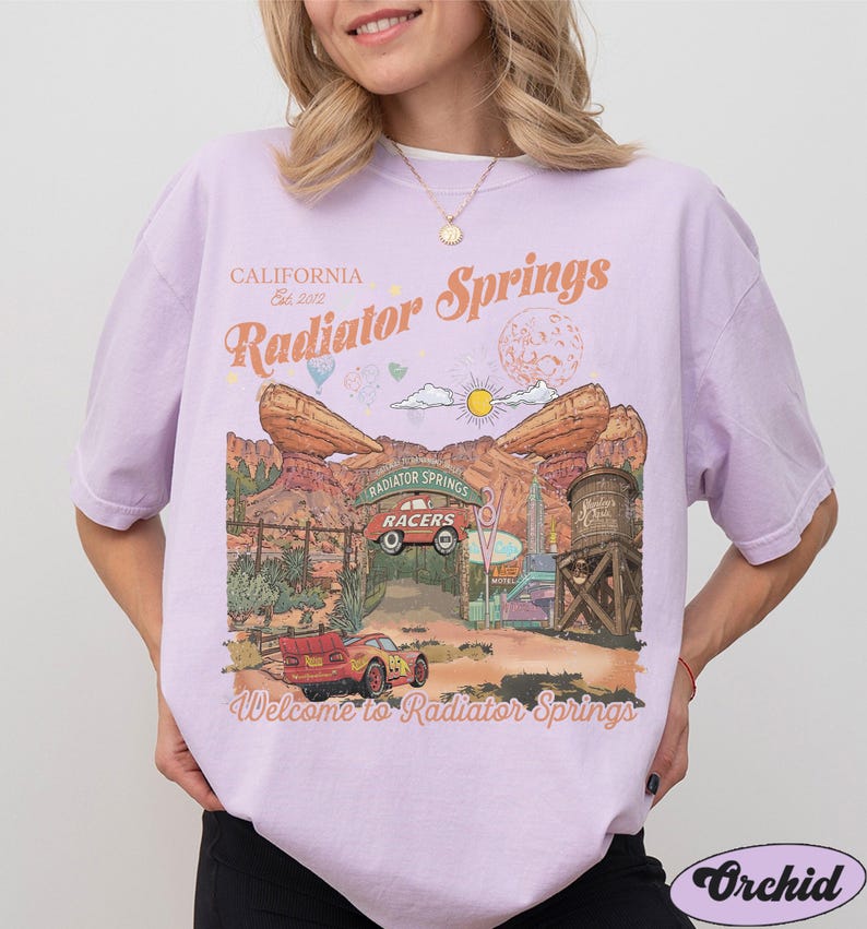 K&ouml;nnte beinhalten: Ein hellviolettes Kurzarm-T-Shirt mit einem Grafikdruck von Radiator Springs und dem Text California Est. 2012, Radiator Springs sowie Welcome to Radiator Springs. Das Design zeigt ein rotes Rennauto, W&uuml;stenfelsformationen und einen Wasserturm.