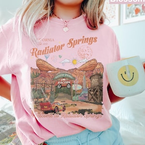 K&ouml;nnte beinhalten: Ein hellrosa T-Shirt mit einem Grafikdruck von Radiator Springs, inklusive des Textes "Radiator Springs" und "Welcome to Radiator Springs". Das Design zeigt ein rotes Rennauto, W&uuml;stenfelsformationen, einen Wasserturm und eine Sonne.