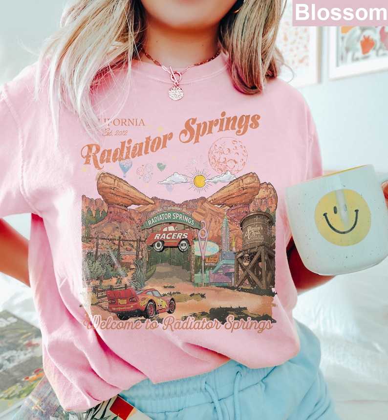 K&ouml;nnte beinhalten: Ein hellrosa T-Shirt mit einem Grafikdruck von Radiator Springs, inklusive des Textes "Radiator Springs" und "Welcome to Radiator Springs". Das Design zeigt ein rotes Rennauto, W&uuml;stenfelsformationen, einen Wasserturm und eine Sonne.