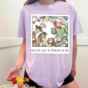 May include: A light purple t-shirt featuring a framed illustration of characters from the movie Toy Story. The illustration includes Woody, Buzz Lightyear, and the Little Green Men. The text "YOU'VE GOT A FRIEND IN ME" is below the illustration.