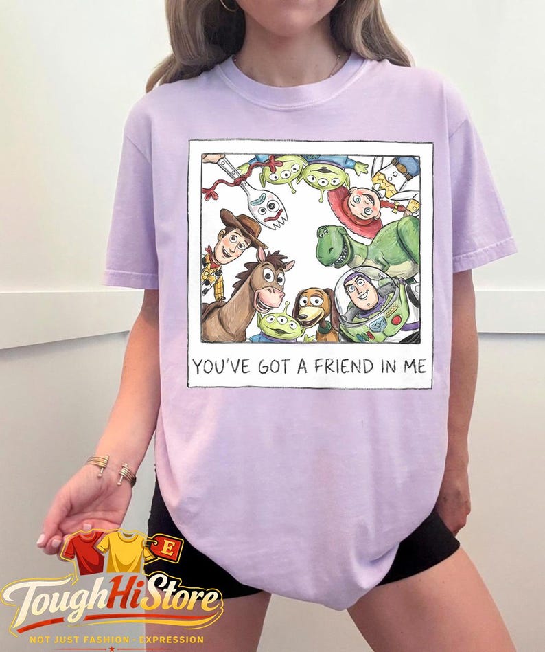 May include: A light purple t-shirt featuring a framed illustration of characters from the movie Toy Story. The illustration includes Woody, Buzz Lightyear, and the Little Green Men. The text "YOU'VE GOT A FRIEND IN ME" is below the illustration.