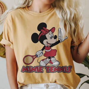 以下が含まれることがあります： テニスウェアを着たミニーマウスが描かれたマスタードイエローのTシャツ。ミニーは赤いドレス、バイザー、靴を身に着け、テニスラケットを持っています。「MINNIE TENNIS」の文字が下に赤色でプリントされています。