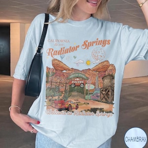 K&ouml;nnte beinhalten: Ein hellblaues T-Shirt mit einem Grafikdruck der Radiator Springs Landschaft, inklusive roter Rennwagen, einem Wasserturm und Felsformationen. Der Text lautet California Est. 2012, Radiator Springs und Welcome to Radiator Springs.