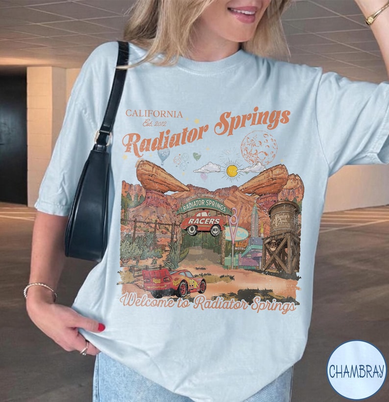 K&ouml;nnte beinhalten: Ein hellblaues T-Shirt mit einem Grafikdruck der Radiator Springs Landschaft, inklusive roter Rennwagen, einem Wasserturm und Felsformationen. Der Text lautet California Est. 2012, Radiator Springs und Welcome to Radiator Springs.
