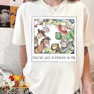 May include: Cream-colored t-shirt featuring a framed watercolor illustration of Toy Story characters. The image includes Woody, Buzz Lightyear, and other characters. The text "YOU'VE GOT A FRIEND IN ME" is printed below the image.