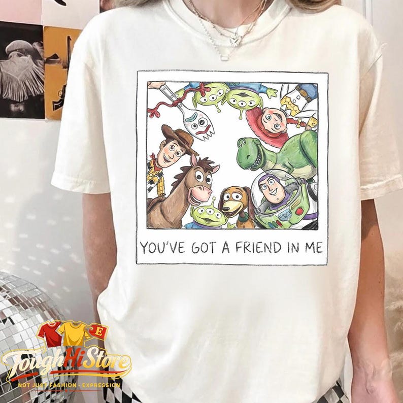 May include: Cream-colored t-shirt featuring a framed watercolor illustration of Toy Story characters. The image includes Woody, Buzz Lightyear, and other characters. The text "YOU'VE GOT A FRIEND IN ME" is printed below the image.
