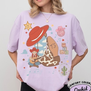 May include: A light purple t-shirt featuring a graphic print of a cowgirl playing a guitar, a red cowboy hat, a cow-print boot with the initials T.S., a disco ball, alphabet blocks, a cactus, musical notes, and a sheriff star labeled Sherf Swift.