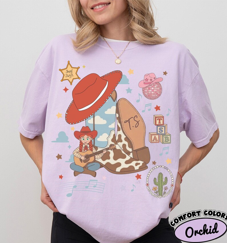 May include: A light purple t-shirt featuring a graphic print of a cowgirl playing a guitar, a red cowboy hat, a cow-print boot with the initials T.S., a disco ball, alphabet blocks, a cactus, musical notes, and a sheriff star labeled Sherf Swift.