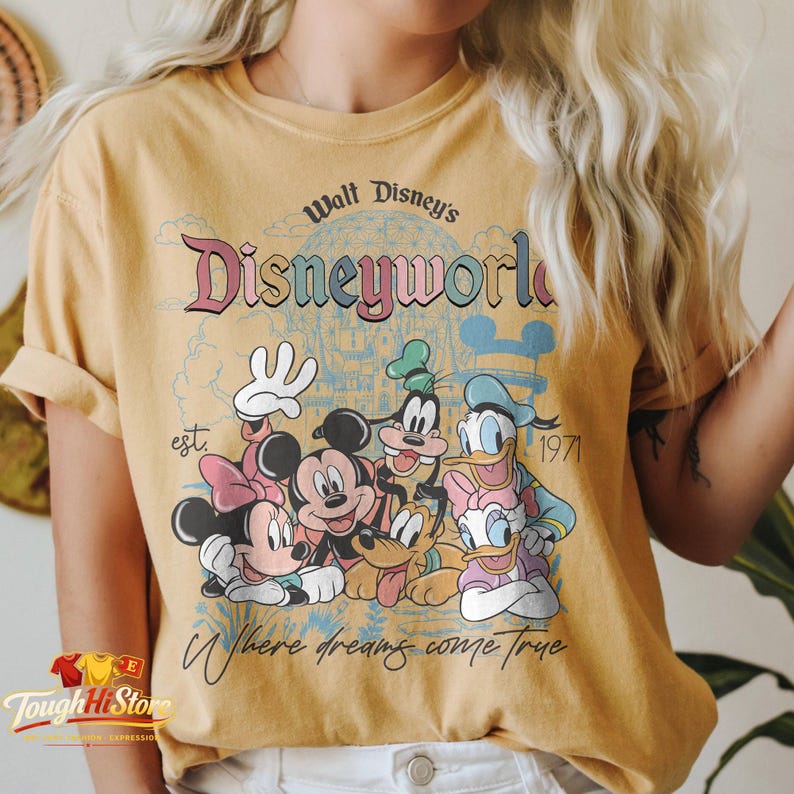 Puede incluir: Camiseta de color amarillo mostaza con un dise&ntilde;o gr&aacute;fico retro de personajes de Disney, incluyendo Mickey, Minnie, Donald, Daisy, Goofy y Pluto. El texto dice "Walt Disney's Disneyworld" y "Where dreams come true".