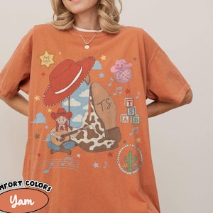 May include: A burnt orange t-shirt featuring a graphic print of a red cowboy hat, a cow-print boot with the letters T.S., a cartoon character playing a guitar, a pink hat, a disco ball, alphabet blocks, a cactus, and a sheriff star labeled Sheriff Swift.