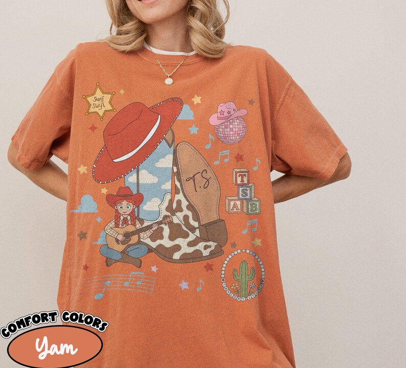 May include: A burnt orange t-shirt featuring a graphic print of a red cowboy hat, a cow-print boot with the letters T.S., a cartoon character playing a guitar, a pink hat, a disco ball, alphabet blocks, a cactus, and a sheriff star labeled Sheriff Swift.