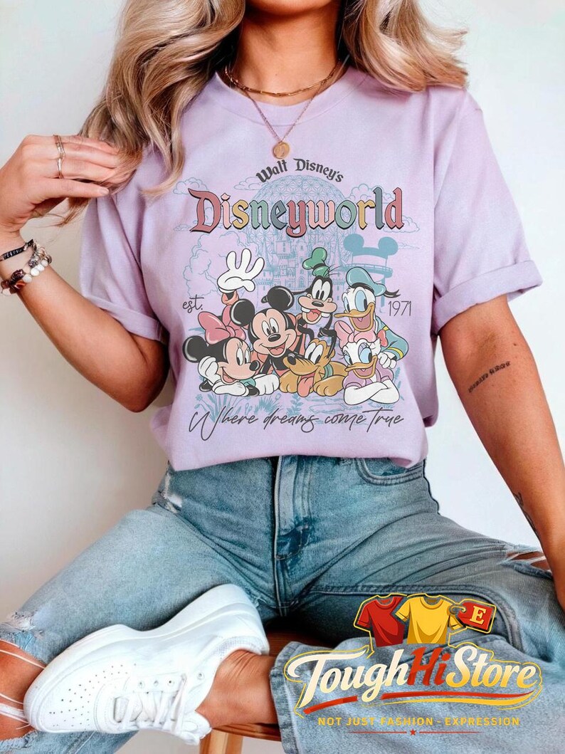 Puede incluir: Camiseta lila claro con un dise&ntilde;o colorido de personajes de Disney y el texto "Walt Disney's Disneyworld" con "est. 1971" y "Where dreams come true". Parte de la colecci&oacute;n Tough Hit Store.