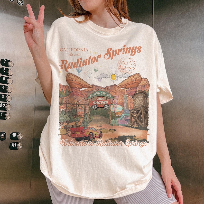 K&ouml;nnte beinhalten: Ein cremefarbenes Kurzarm-T-Shirt mit einem Grafikdruck von Radiator Springs in Kalifornien. Das Design zeigt W&uuml;stenfelsformationen, ein rotes Rennauto, einen Wasserturm sowie den Schriftzug "Radiator Springs" und "Welcome to Radiator Springs".