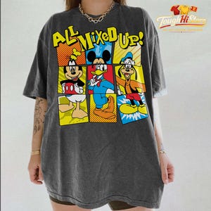 Puede incluir: Camiseta gris oscuro con un gr&aacute;fico colorido de Mickey Mouse, el Pato Donald y Goofy. El texto "ALL MIXED UP!" se muestra encima de los personajes en amarillo y azul. La camiseta tiene un aspecto vintage y desgastado.
