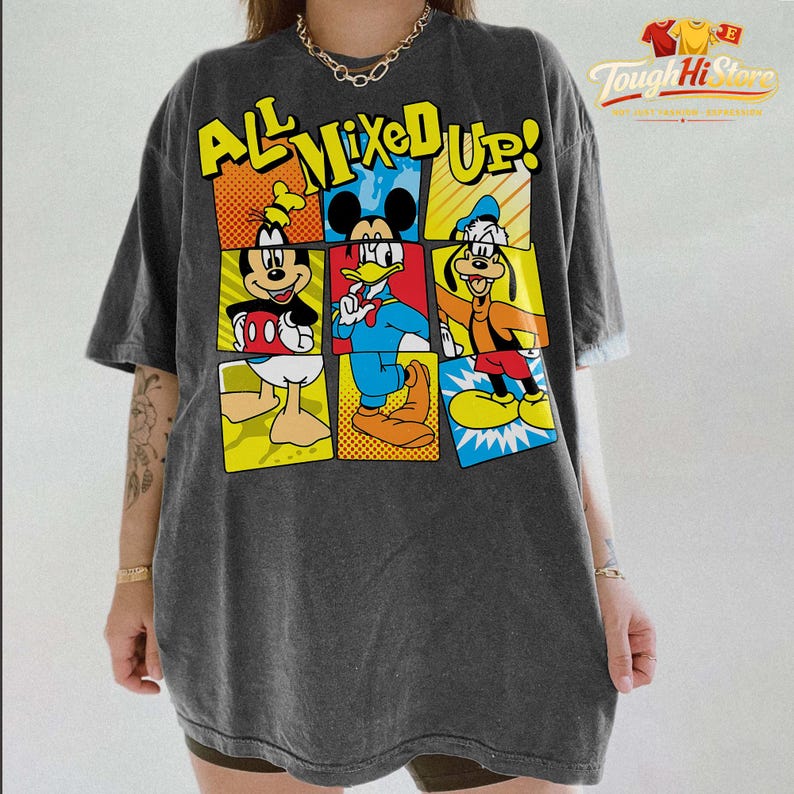 Puede incluir: Camiseta gris oscuro con un gr&aacute;fico colorido de Mickey Mouse, el Pato Donald y Goofy. El texto "ALL MIXED UP!" se muestra encima de los personajes en amarillo y azul. La camiseta tiene un aspecto vintage y desgastado.