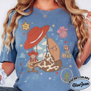 May include: A blue t-shirt featuring a graphic print of a cowgirl playing a guitar, a large red cowboy hat, a cow-print boot with the initials T.S., a sheriff star, a disco ball, alphabet blocks spelling TSB, musical notes, and a cactus.