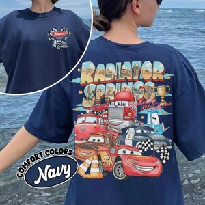 May include: Navy blue t-shirt featuring a graphic of various cartoon cars with the text "Radiator Springs" on the back. The front of the shirt has a small car graphic and the word "Name". The shirt also has the words "Comfort Colors Navy".