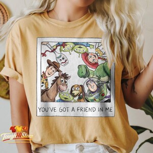May include: Mustard yellow t-shirt featuring a watercolor-style image of Toy Story characters. The image includes Woody, Buzz Lightyear, Rex, and others, framed with the text "YOU'VE GOT A FRIEND IN ME".