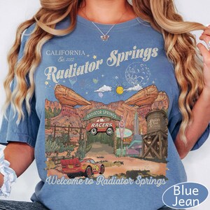 K&ouml;nnte beinhalten: Ein blaues T-Shirt mit kurzen &Auml;rmeln und einem Grafikdruck von Radiator Springs, der ein rotes Rennauto, W&uuml;stenfelsen, einen Wasserturm sowie den Text "California Est. 2012 Radiator Springs" und "Welcome to Radiator Springs" zeigt.