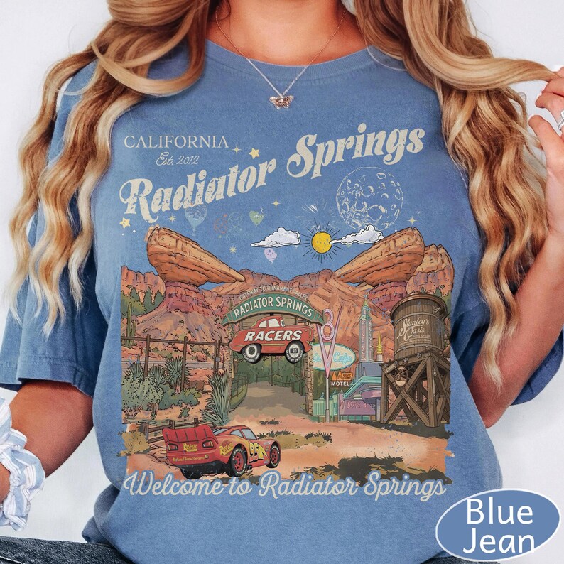 K&ouml;nnte beinhalten: Ein blaues T-Shirt mit kurzen &Auml;rmeln und einem Grafikdruck von Radiator Springs, der ein rotes Rennauto, W&uuml;stenfelsen, einen Wasserturm sowie den Text "California Est. 2012 Radiator Springs" und "Welcome to Radiator Springs" zeigt.