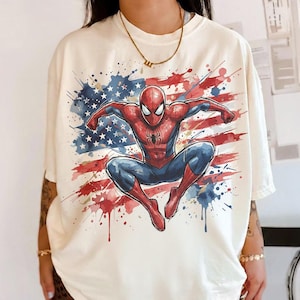 Puede incluir: Camiseta color crema con una vibrante ilustración de Spider-Man en una pose dinámica. El superhéroe se encuentra sobre un fondo de la bandera estadounidense, con salpicaduras de pintura roja, blanca y azul. La camiseta tiene mangas cortas.
