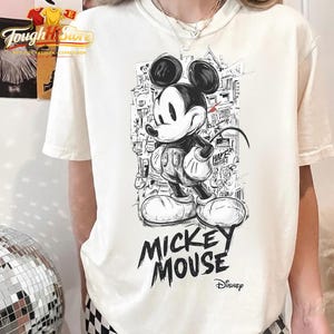 May include: A cream-coloured t-shirt with a black and white Mickey Mouse illustration in a street art style. The words "MICKEY MOUSE" are printed in a bold, graffiti-like font below the character. The Disney logo is also visible.