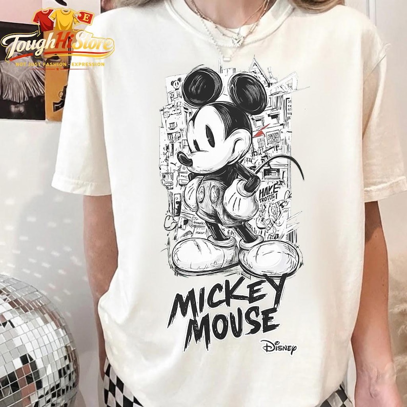 May include: A cream-coloured t-shirt with a black and white Mickey Mouse illustration in a street art style. The words "MICKEY MOUSE" are printed in a bold, graffiti-like font below the character. The Disney logo is also visible.