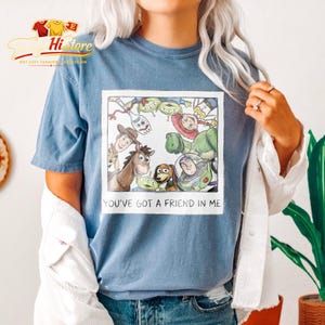 May include: Blue t-shirt featuring a framed graphic of Toy Story characters, including Woody, Buzz Lightyear, and Rex. The text "YOU'VE GOT A FRIEND IN ME" is printed below the image. The shirt has short sleeves.