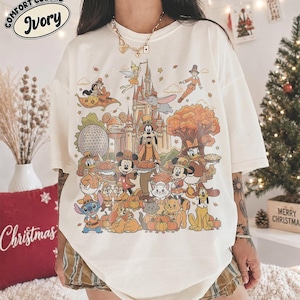 Puede incluir: Una camiseta de color marfil con un estampado gráfico colorido de varios personajes de Disney, incluidos Mickey Mouse, Goofy y Campanilla, frente a un castillo. El texto "Comfort Colors" e "Ivory" aparece en la esquina superior izquierda.